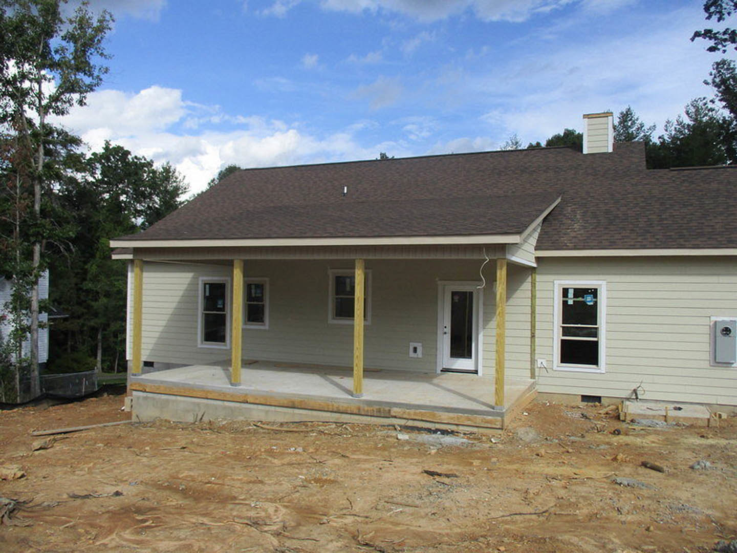 Large porch with unfinished wood framing, white entry door with glass panel, window displaying construction sign, exposed siding, dirt ground, and nearby tree under cloudy sky.