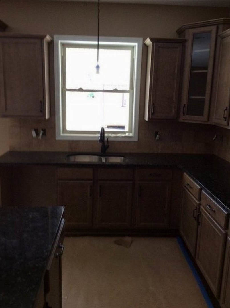 Modern kitchen featuring white cabinetry, light stone countertops, stainless steel sink beneath a large window, black dining table in foreground, wood flooring, and minimalist