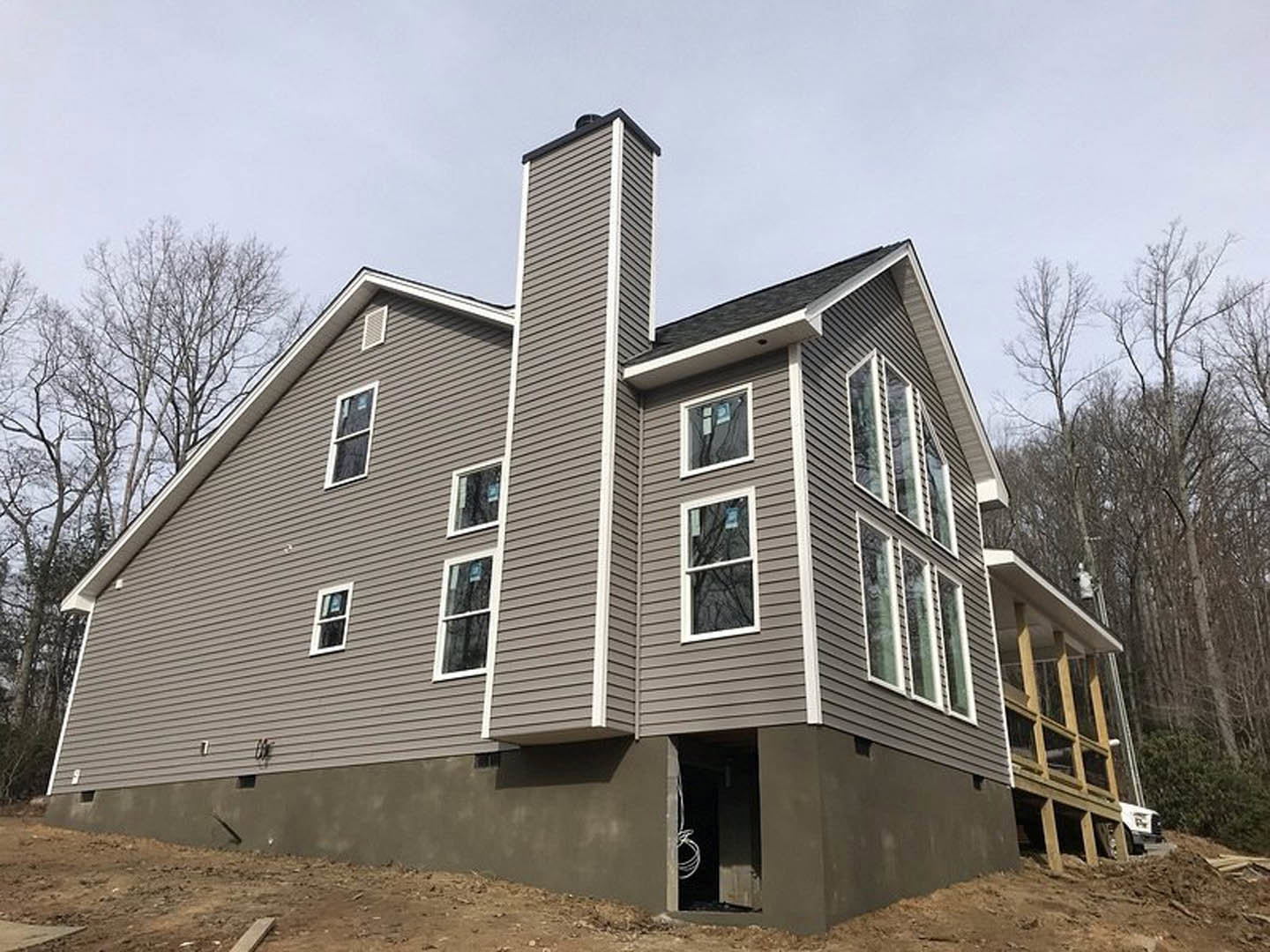 Wood-framed house under construction with covered porch, white-trimmed window, exposed chimney, unfinished siding, and roof surrounded by trees