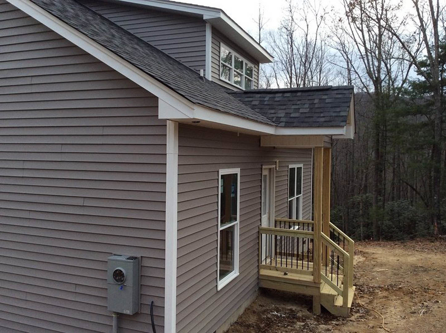 Wooden stairs with railing lead to a covered porch, light-colored siding, large window, and circular wall fixture; dirt area in foreground, tree branches visible above.