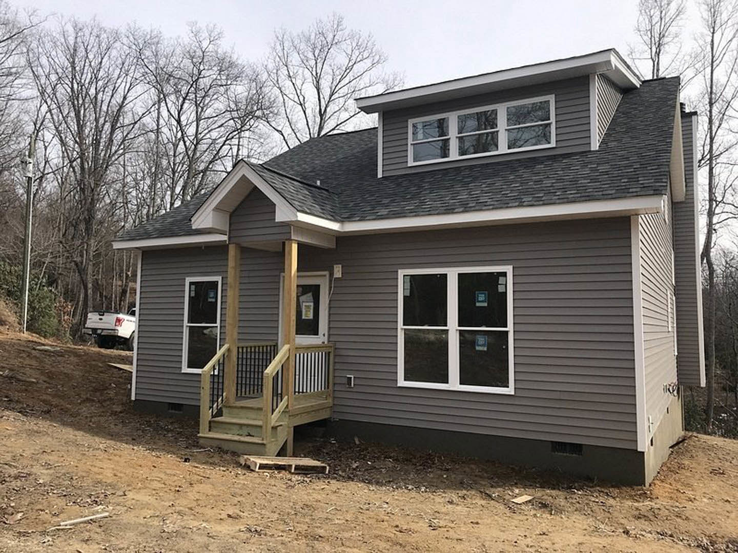 Two-story cottage with light siding, covered porch, wooden stairs, white-framed windows, metal support post, and a white truck parked among trees.