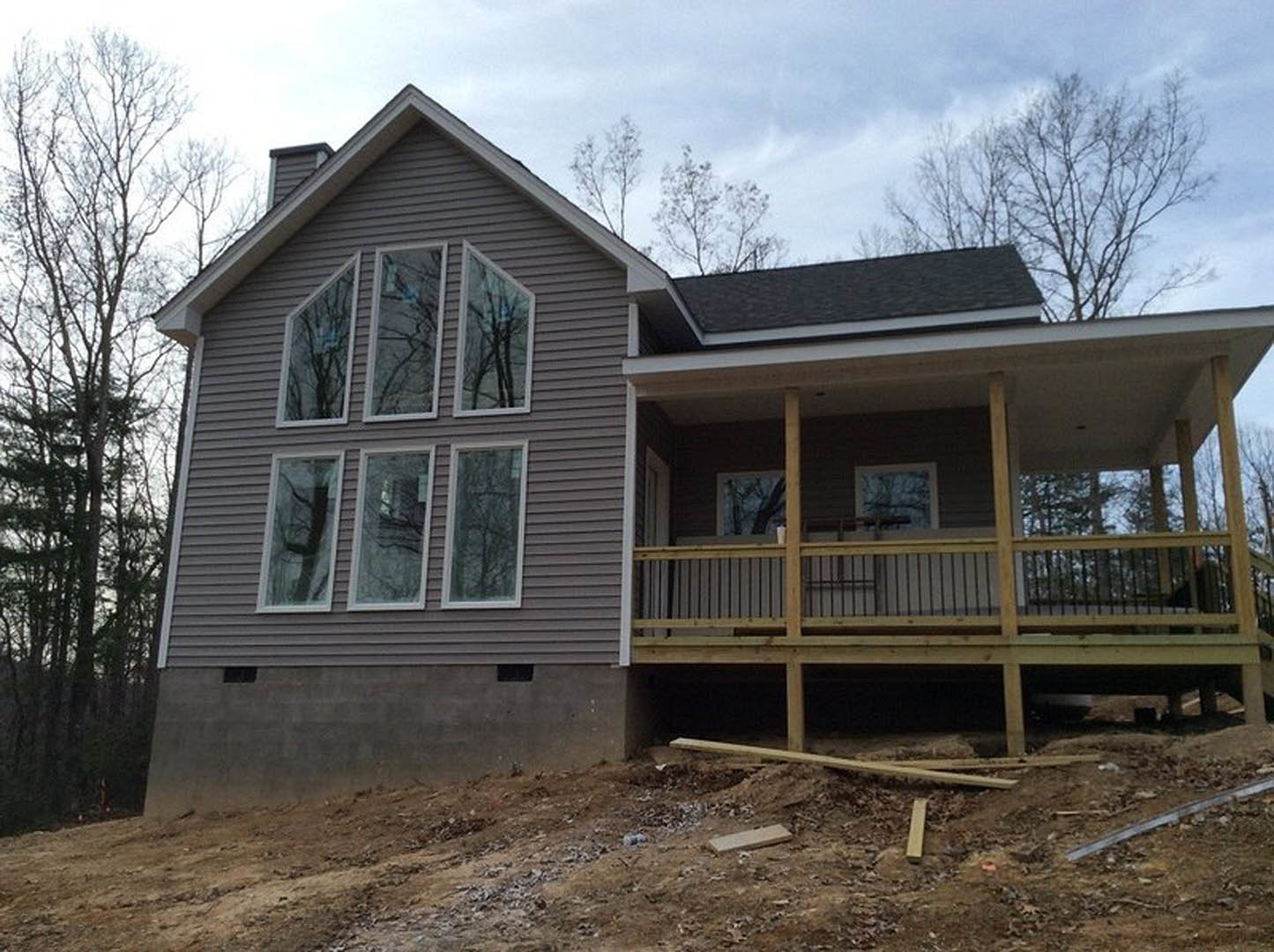 Partially built house with exposed wooden deck and railing, white-framed windows, gray siding, tree branches near window, and unfinished roof under blue sky