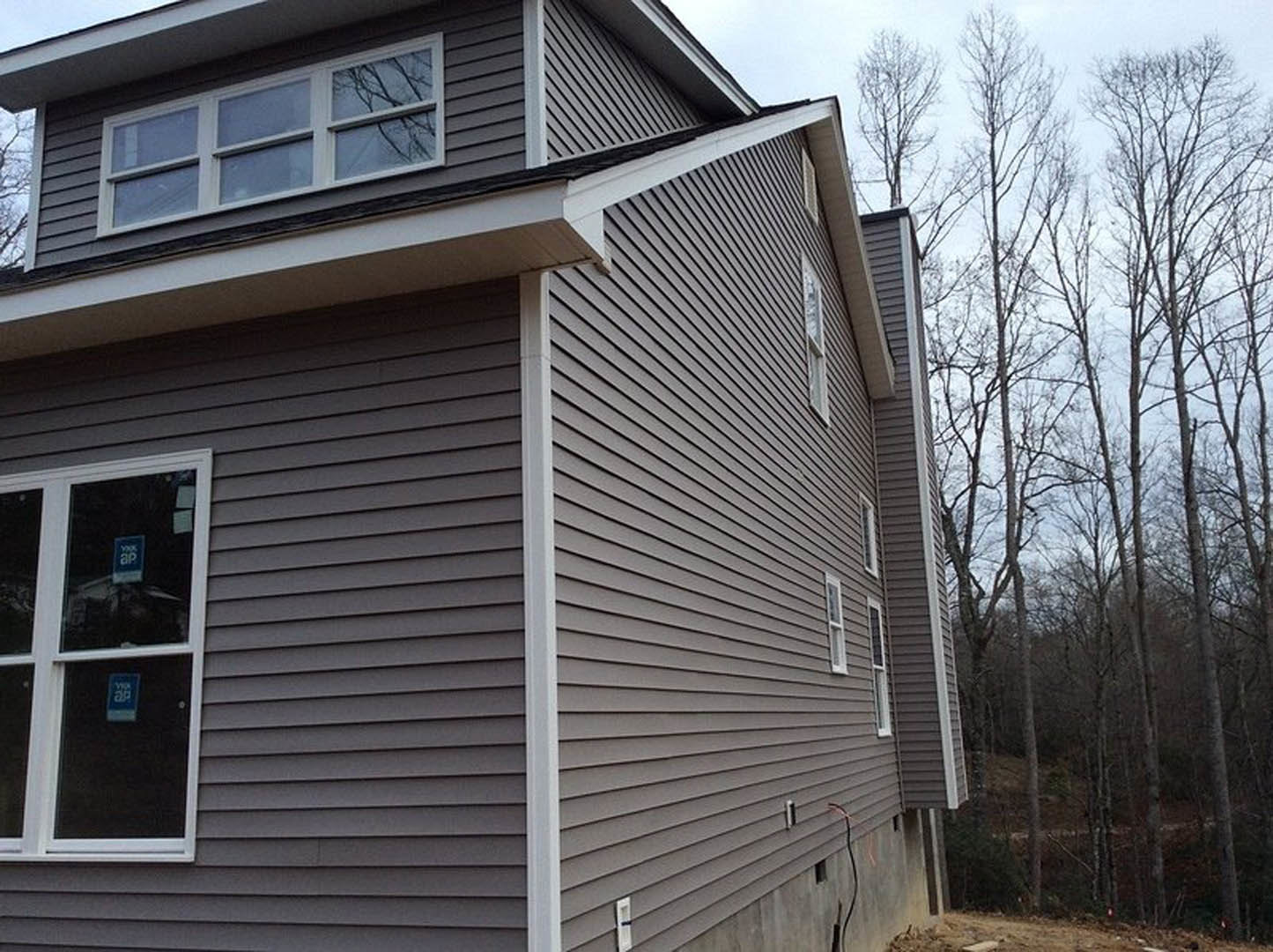 Two-story cottage with light siding, dark roof, and minimal windows, surrounded by mature trees; blue sign with white text displayed in front window.