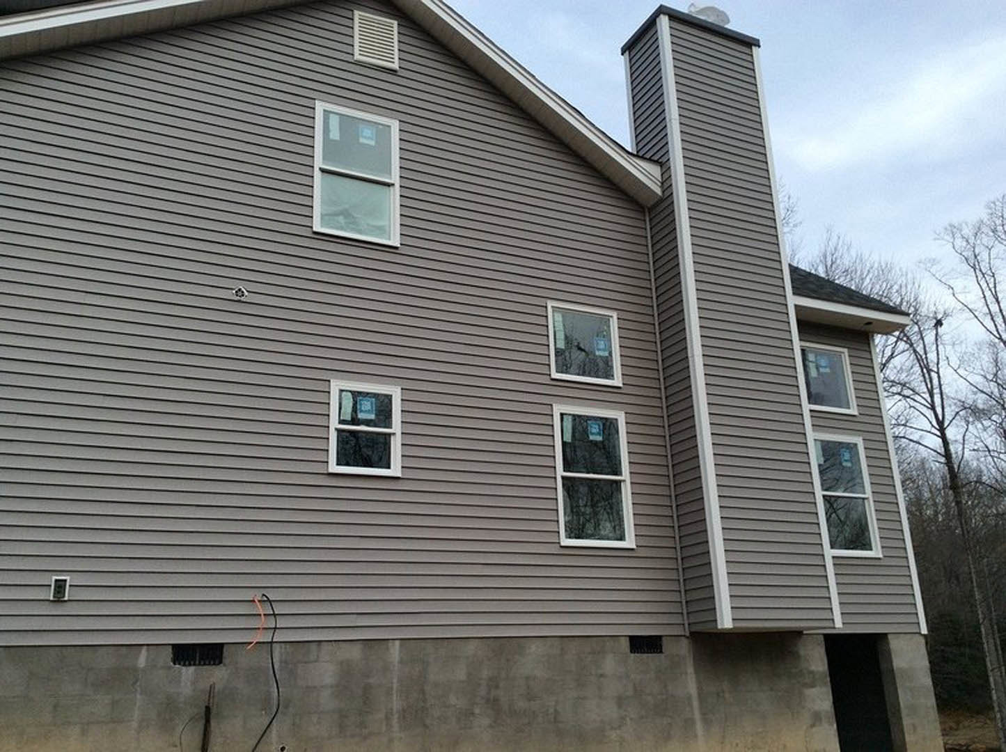 Two-story home with gray siding, multiple large white-framed windows, brick chimney, and gable roof surrounded by trees.