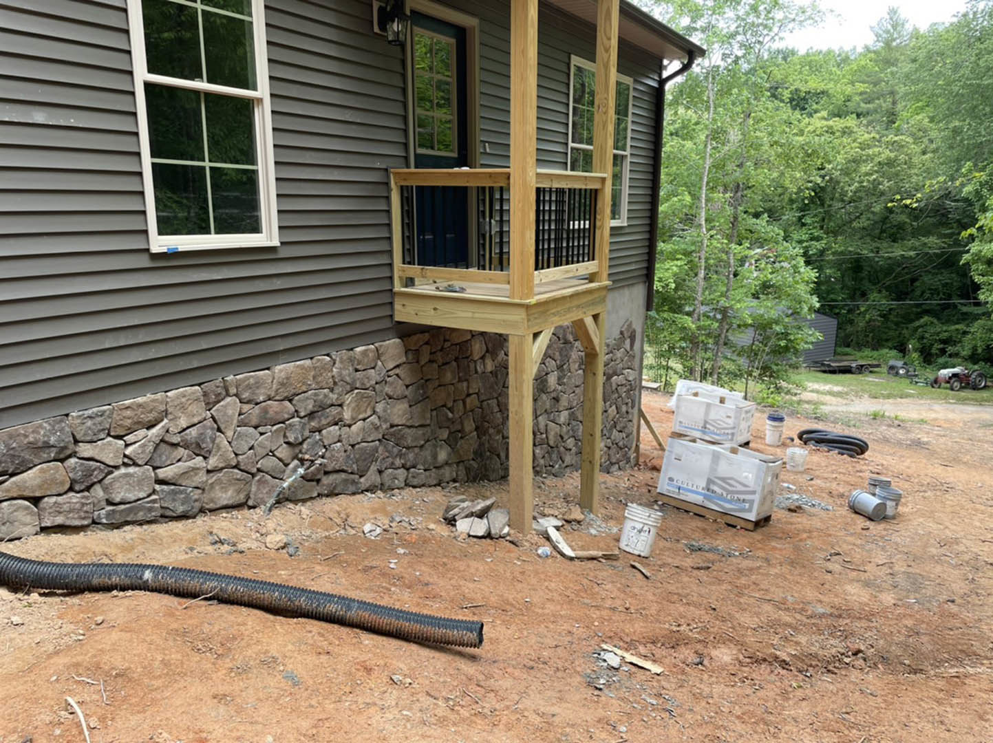 Front porch with wooden decking, stone wall facade, large window, white utility box on pallet, black pipe and white bucket on ground, surrounded by trees and plants