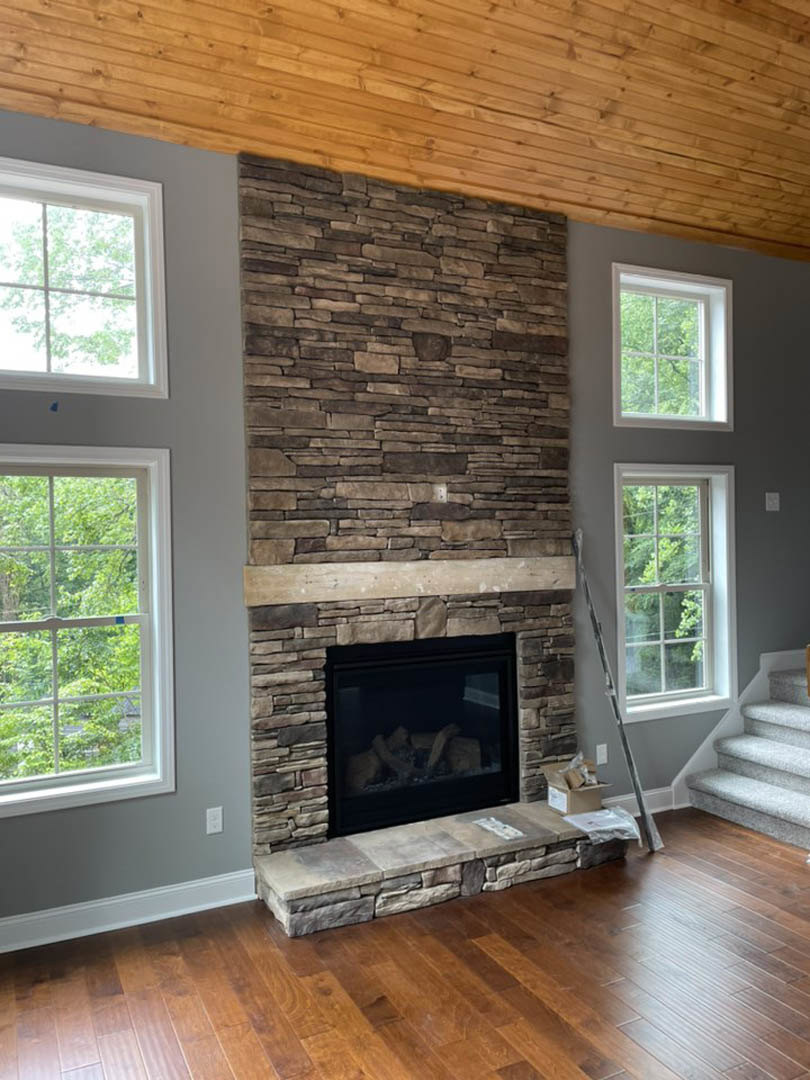Stone fireplace with glass door and wood beam mantel, adjacent to window with white frame and blue tape, wood flooring and stone step in cozy den.
