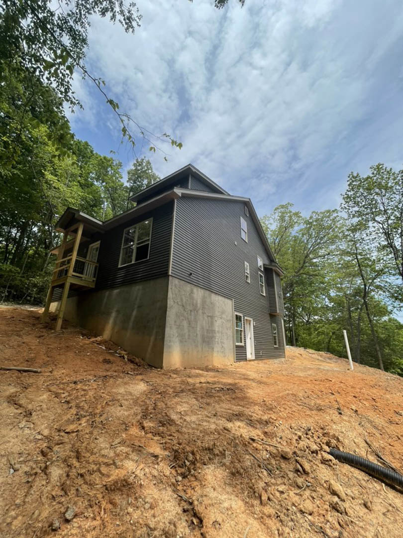 Wood-framed house under construction on a dirt lot surrounded by tall trees, with exposed balcony and staircase facing a wooded hillside