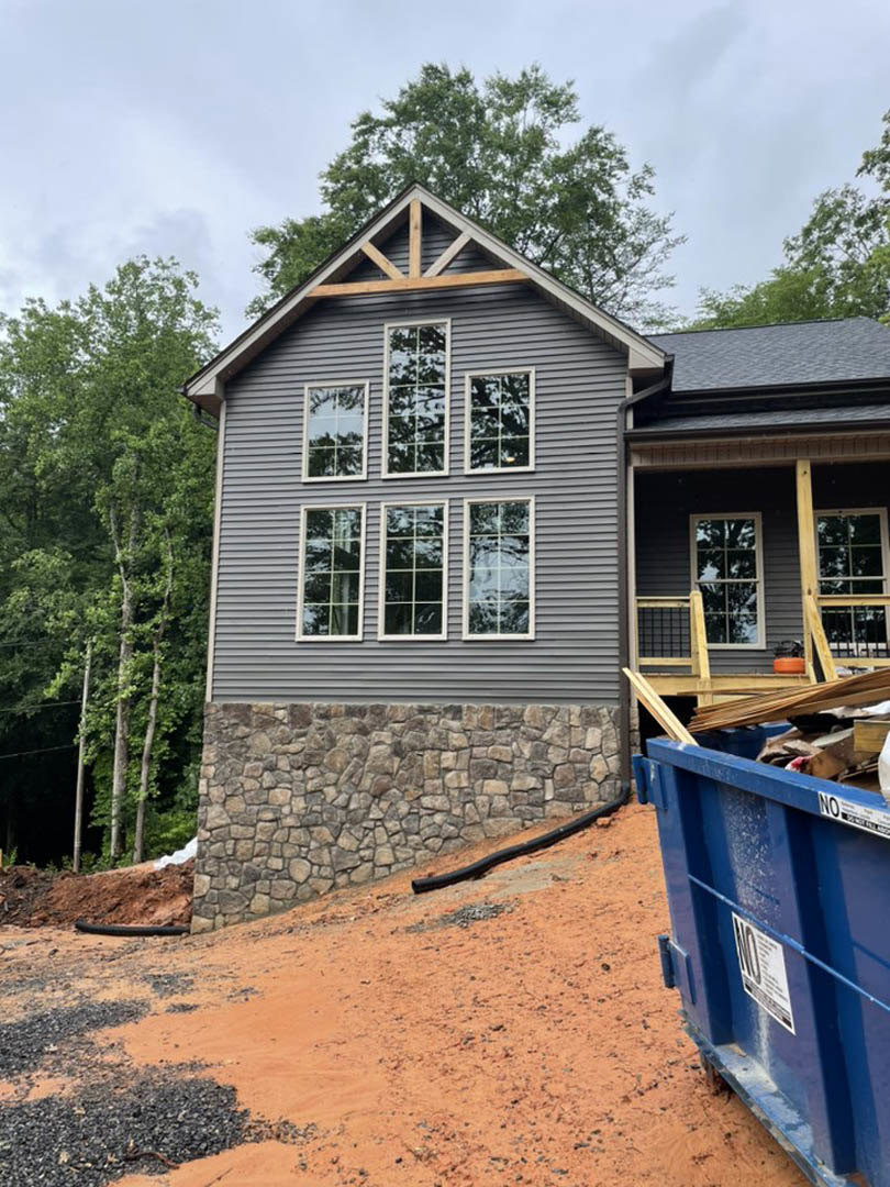 White-framed windows with tree reflections on light siding, blue waste container with white label, black pipe, and pile of trash scattered on ground in front of cottage-style home