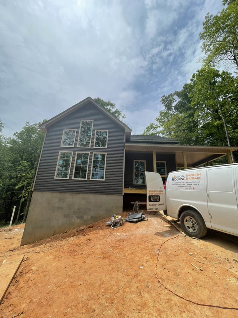 Modern two-story home with multiple large windows, light-colored siding, and a white van parked on a dirt driveway; fenced yard and construction equipment visible in foreground