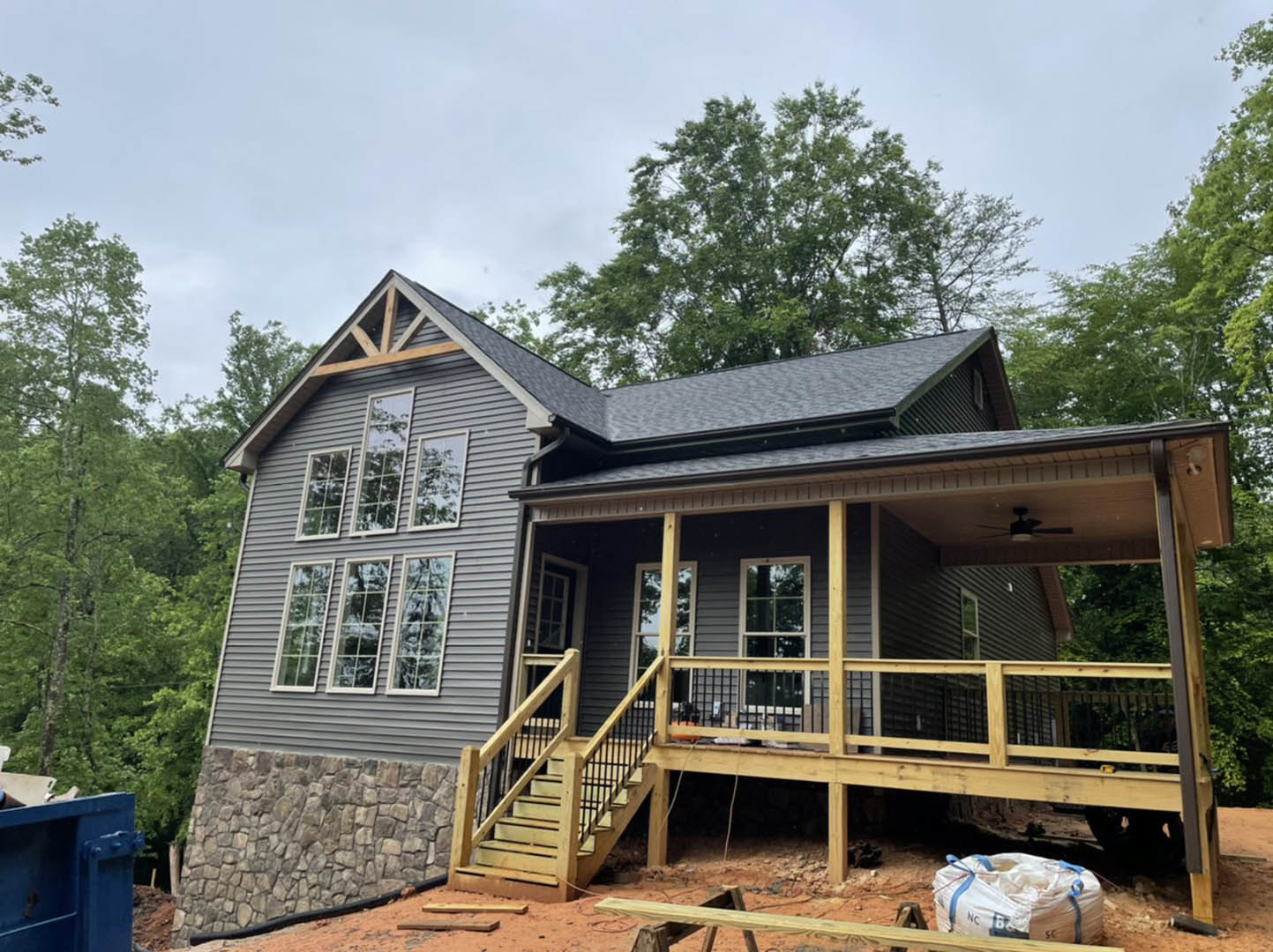 Wood deck under construction with exposed framing, wooden stairs leading to unfinished entry, stone wall featuring black accent band, blue metal waste container with open door