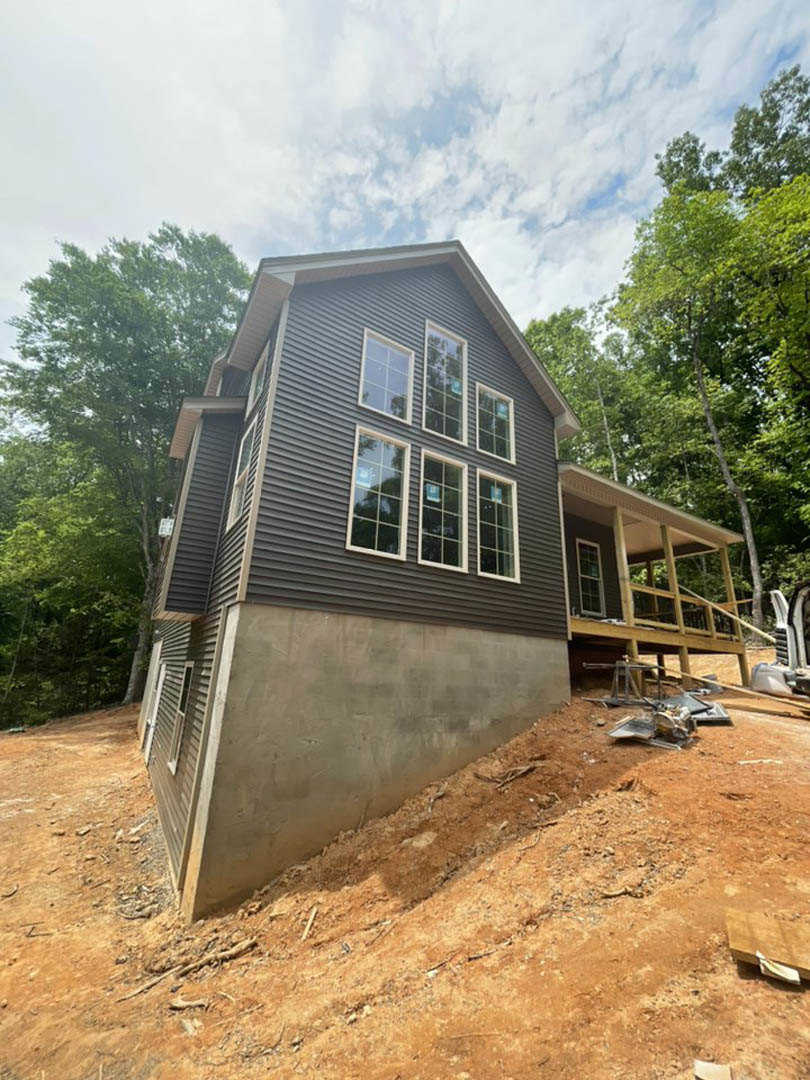 Partially built house with exposed framing, white-trimmed square pane window, sloped dirt hill in foreground, mature trees and cloudy sky in background