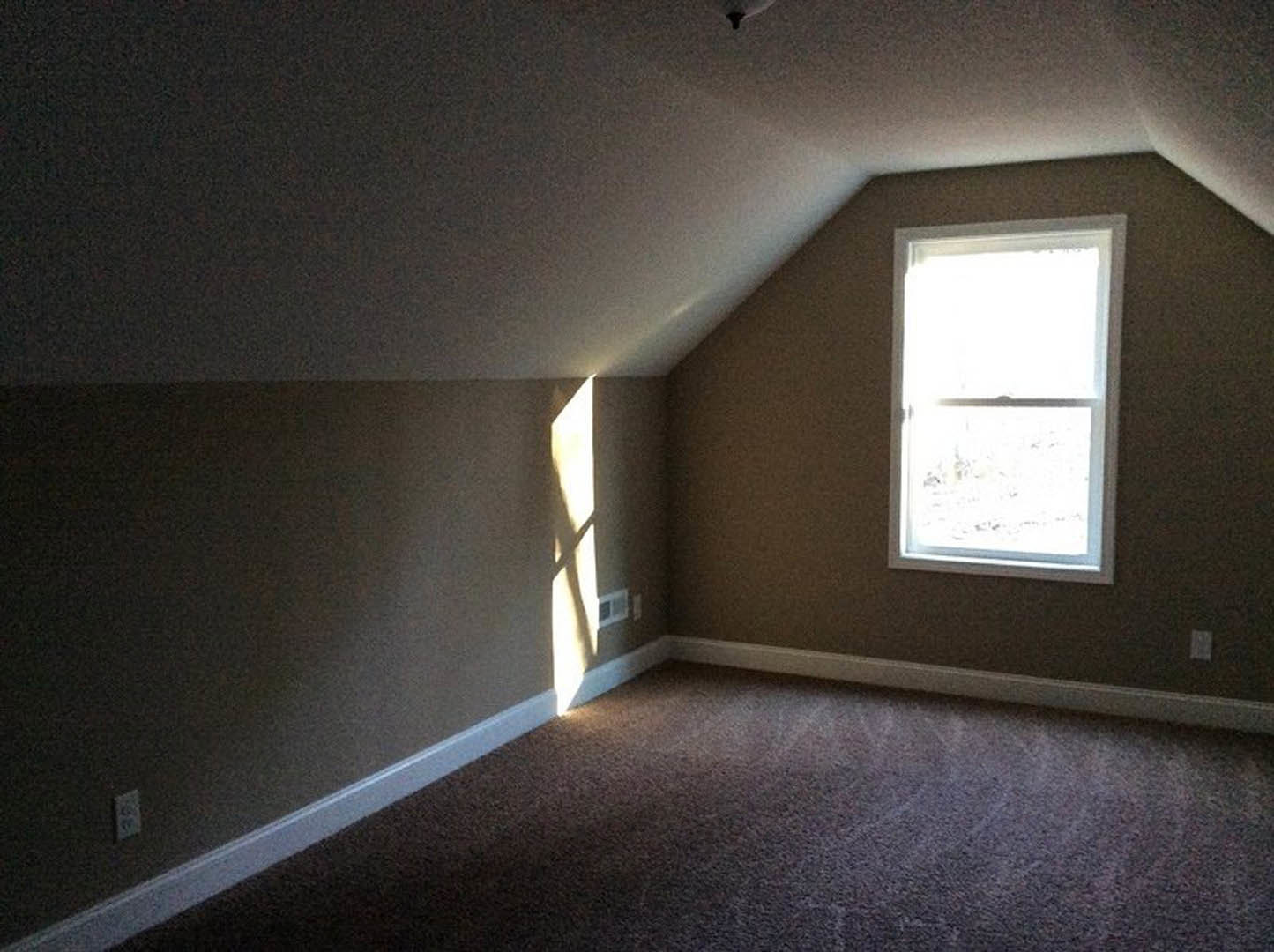 Carpeted floor with white baseboard trim, white electrical outlet, large window with white frame and vent, plaster walls