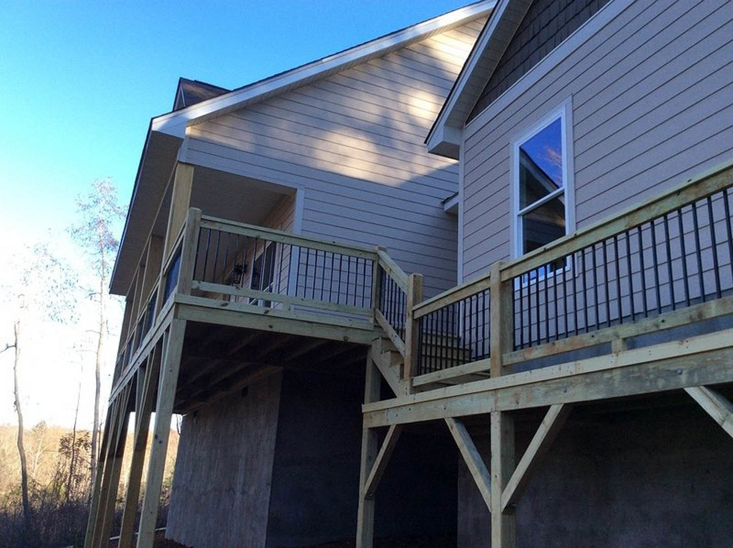 Wooden deck with horizontal railing, exposed beams, large window reflecting blue sky, tree branches nearby, light-colored siding exterior