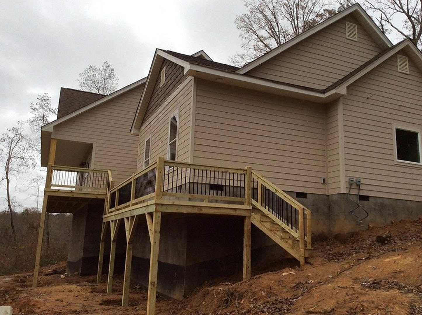 Two-story house with light siding, wooden deck featuring railing and stairs, large window with dark trim, leafless tree in foreground, blue sky above