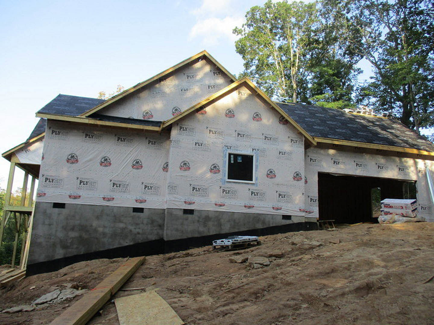 Partially built house wrapped in a large white protective cover, exposed framing visible, construction materials scattered on bare ground, trees and sky in background
