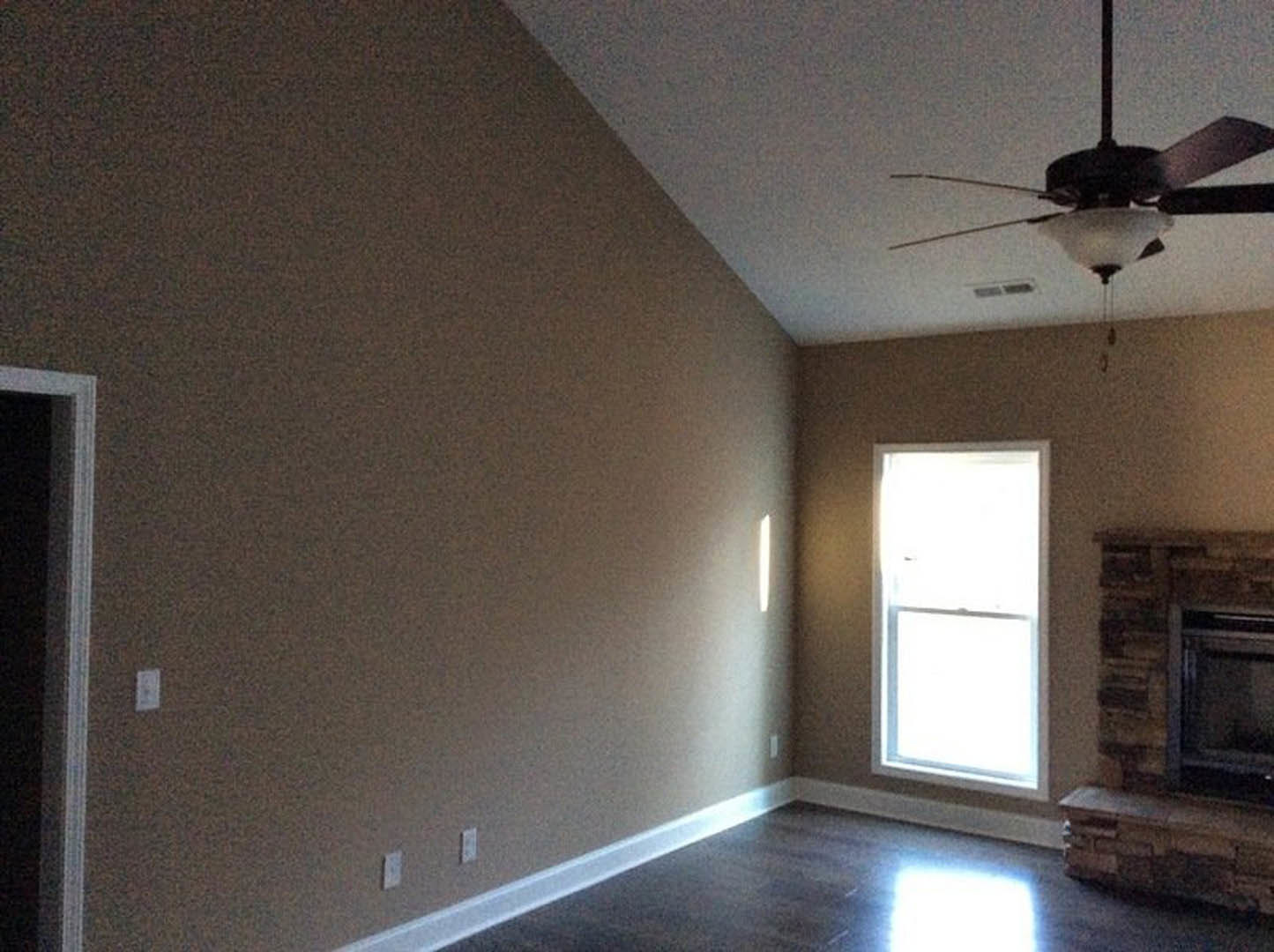Ceiling fan with light fixture mounted on white ceiling, sunlight streaming through window onto light-colored floor, partial view of fireplace and neutral walls.