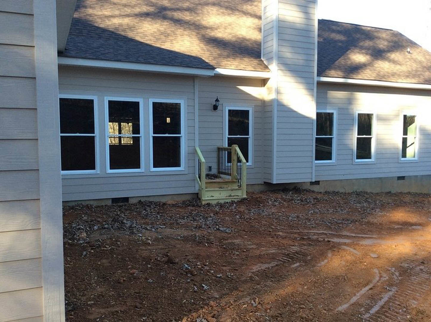 Two-story house with light siding, multiple windows with white frames, wooden stairs featuring metal railing, dirt ground with visible tire tracks in front