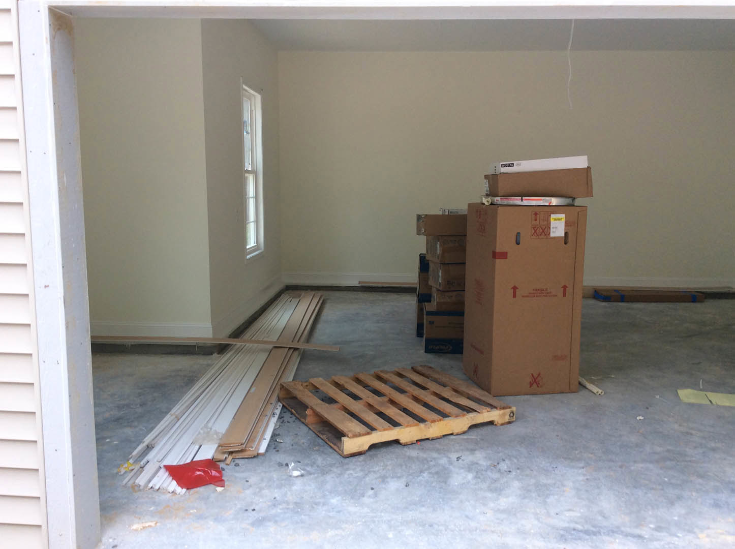 Concrete floor with stacked wooden pallets, cardboard boxes with red labels, white-framed window, pile of wood planks, small red item on the ground