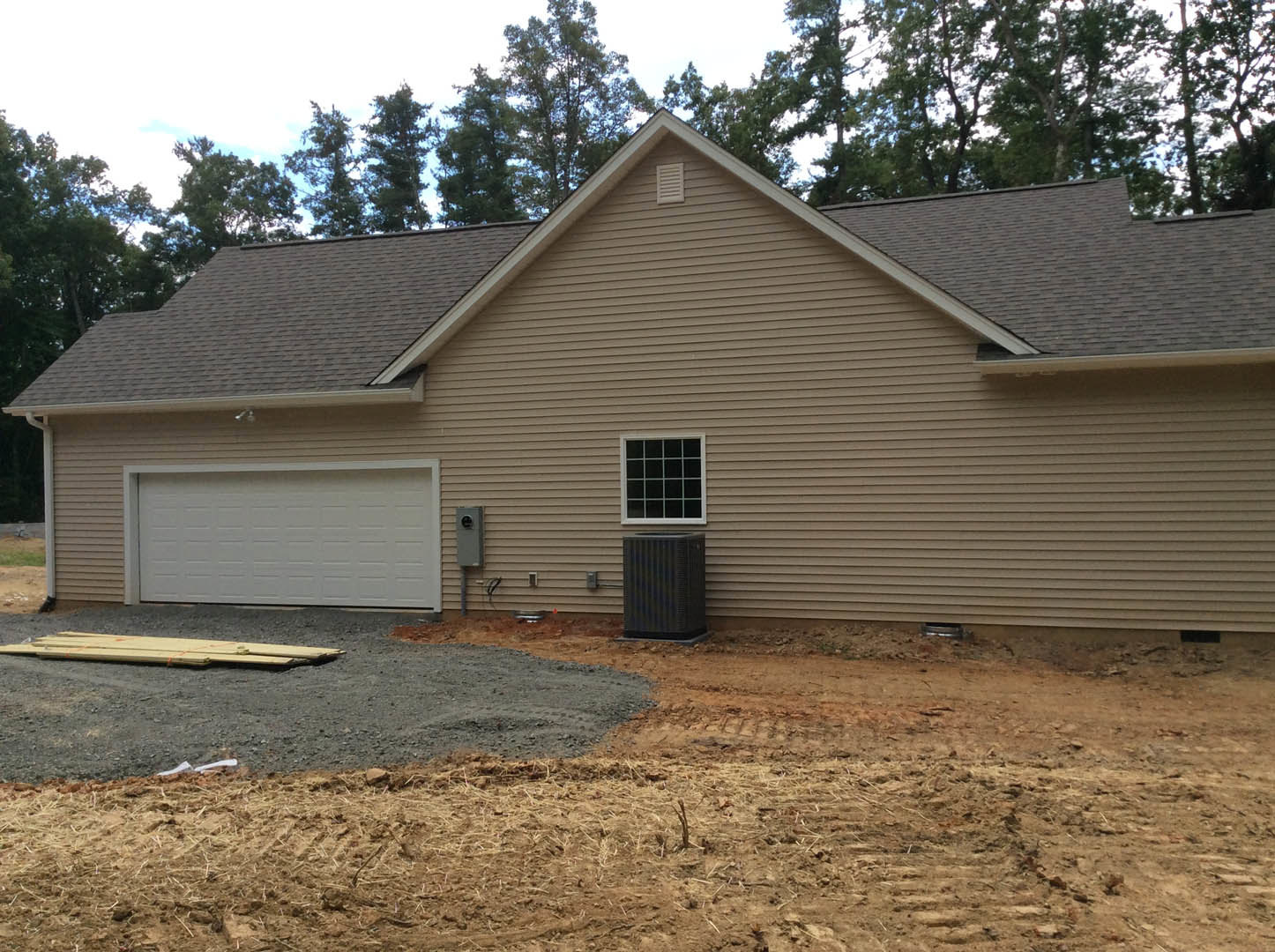 White garage door with matching trim, gravel driveway leading to entry, light-colored siding, multi-pane windows, stack of wood planks near house, trees in background.