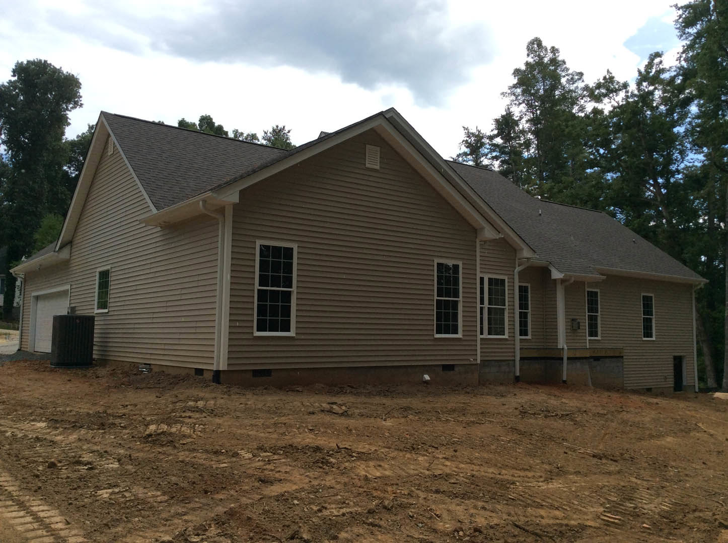 Two-story house under construction with white siding, large multi-pane windows, dirt yard, and surrounding trees