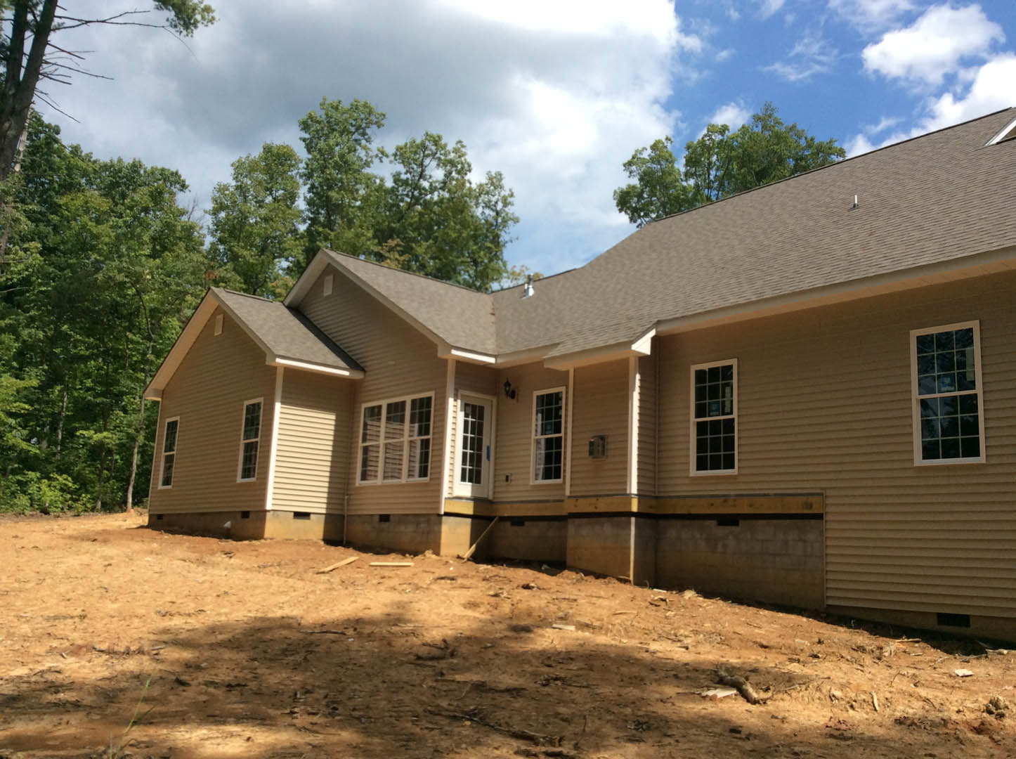 Two-story house under construction with exposed framing, multiple square-paned windows, dirt yard, and mature trees in the background