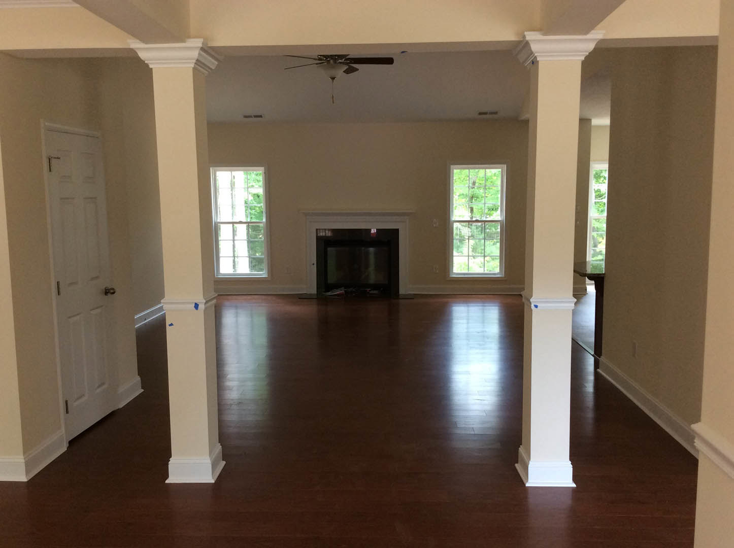 Living room with white columns, built-in fireplace with mounted TV, hardwood floors, large windows framed in white showing tree views, and a white door with silver hardware.