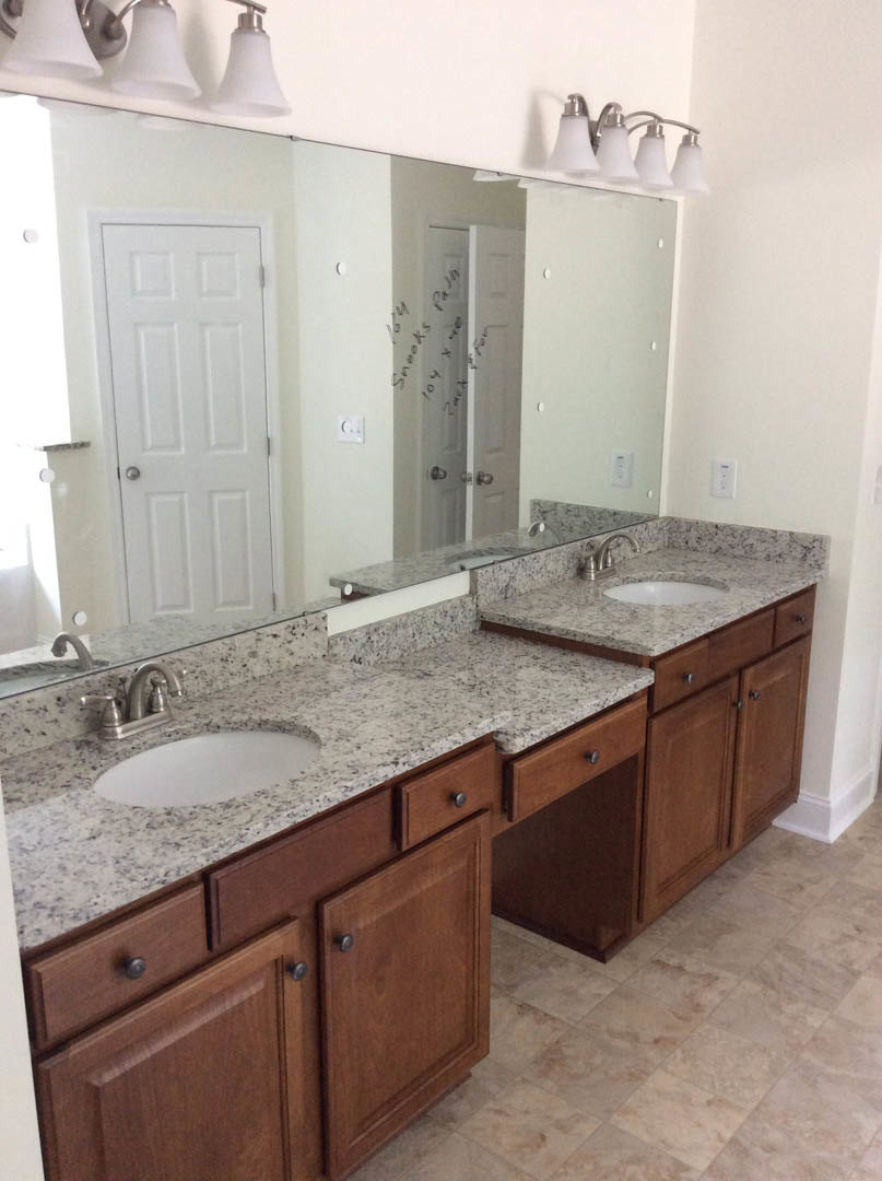 Bathroom with double sinks set in a stone countertop, large mirror above, wooden cabinetry below, silver faucets, white door with silver handle, and three-light fixture mounted on
