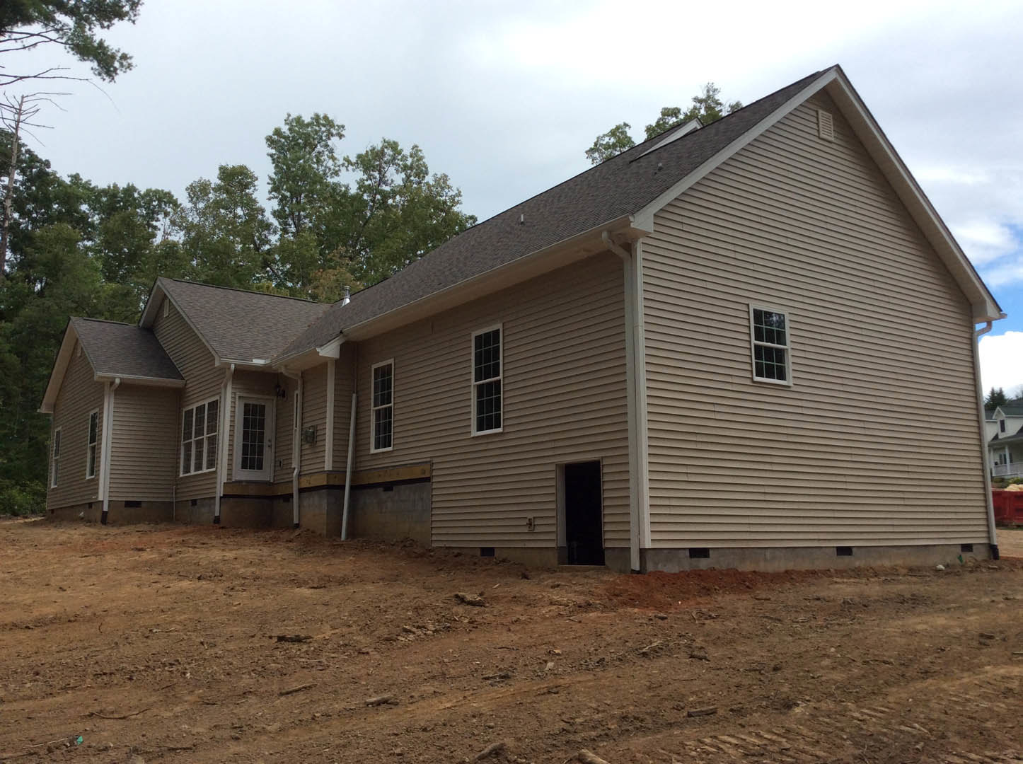 Two-story house under construction with white-framed windows, light siding, exposed dirt yard, and surrounding trees