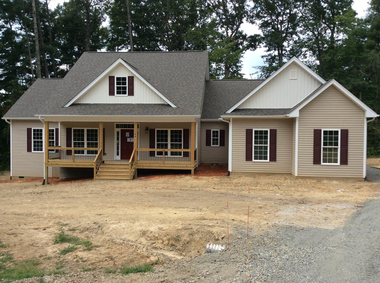 Partially built house with white-trimmed windows, covered porch, exterior door displaying papers, unfinished roof, surrounding trees, and dirt excavation with exposed metal pipe