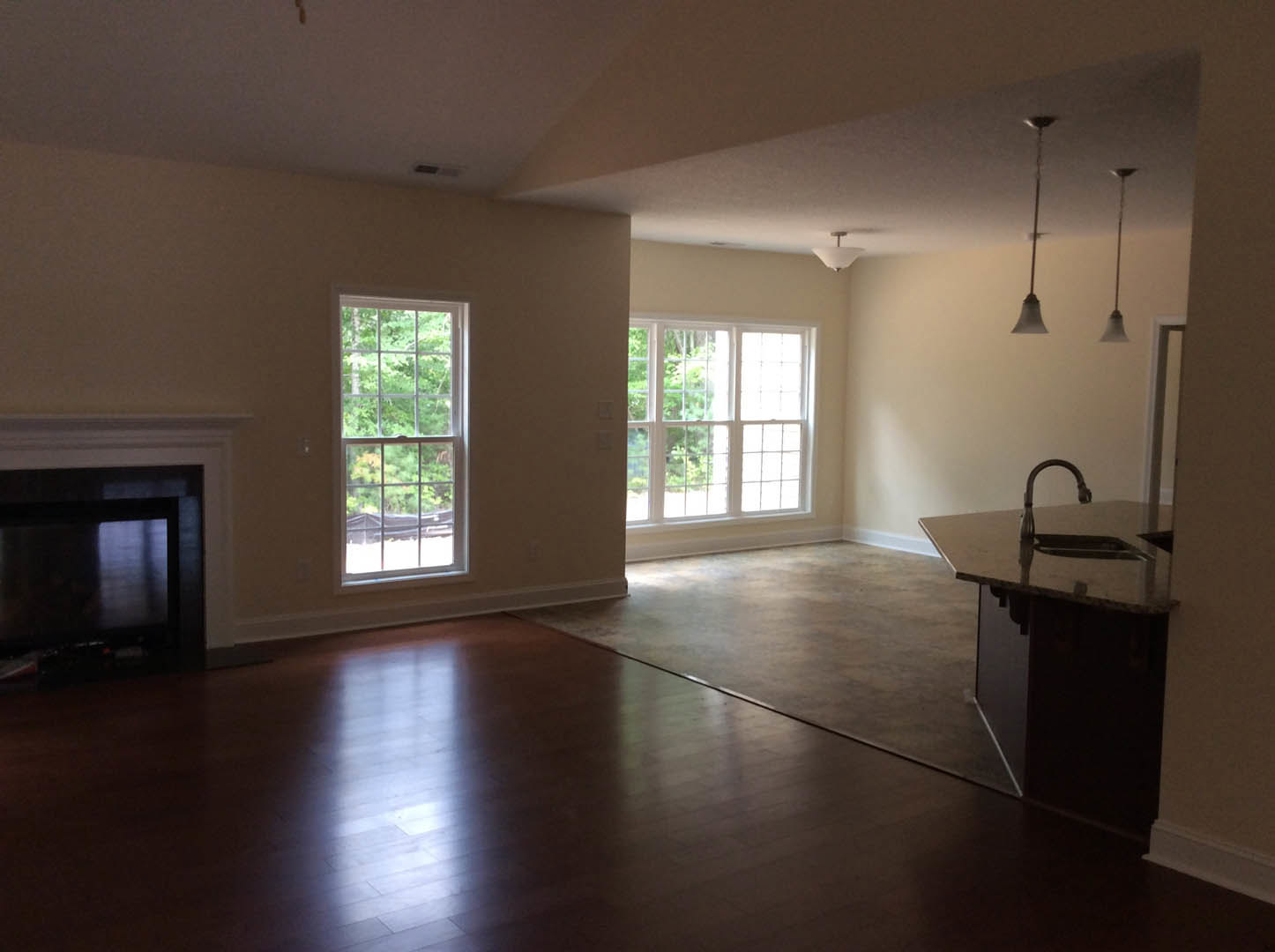 Open-concept living room with wood flooring, white fireplace, large multi-pane window overlooking trees, and adjacent kitchen area