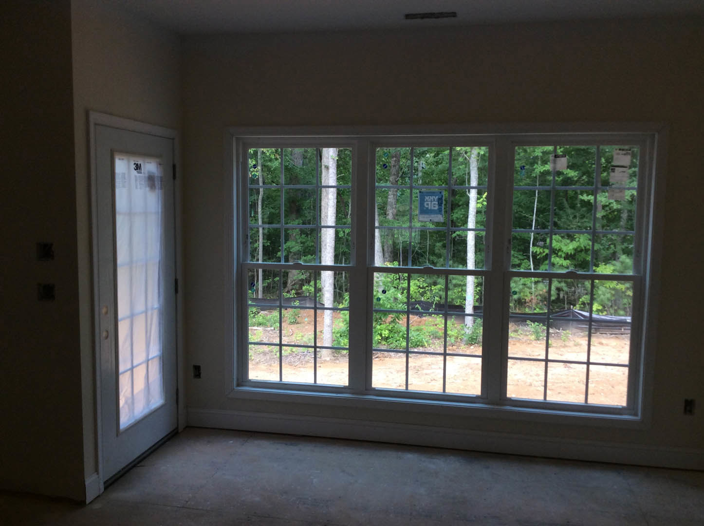 Bright room with large windows overlooking trees, white walls, grey flooring, and a modern door with blue sign