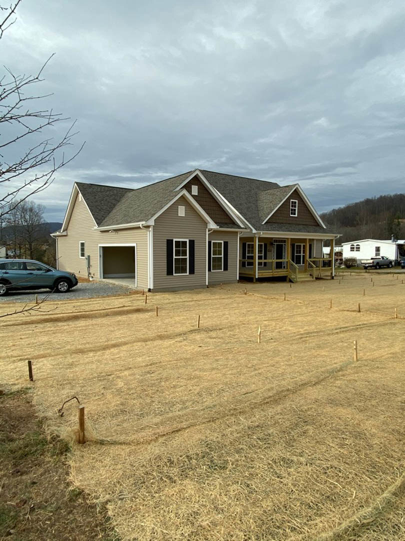 Two-story custom home with white garage door, large windows, gray siding, and a parked car on the driveway; manicured lawn, tree branches, and blue sky in the background.