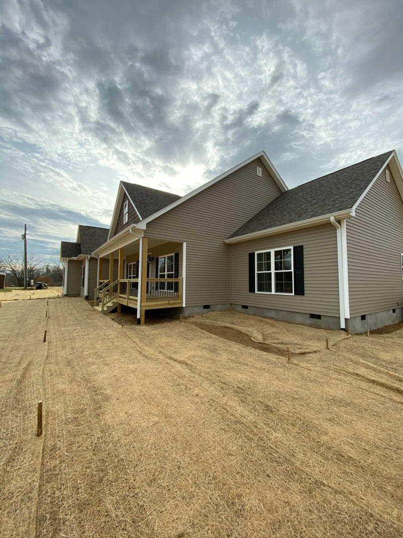 Framed house under construction with exposed wood, unfinished porch, large windows, and cloudy sky overhead