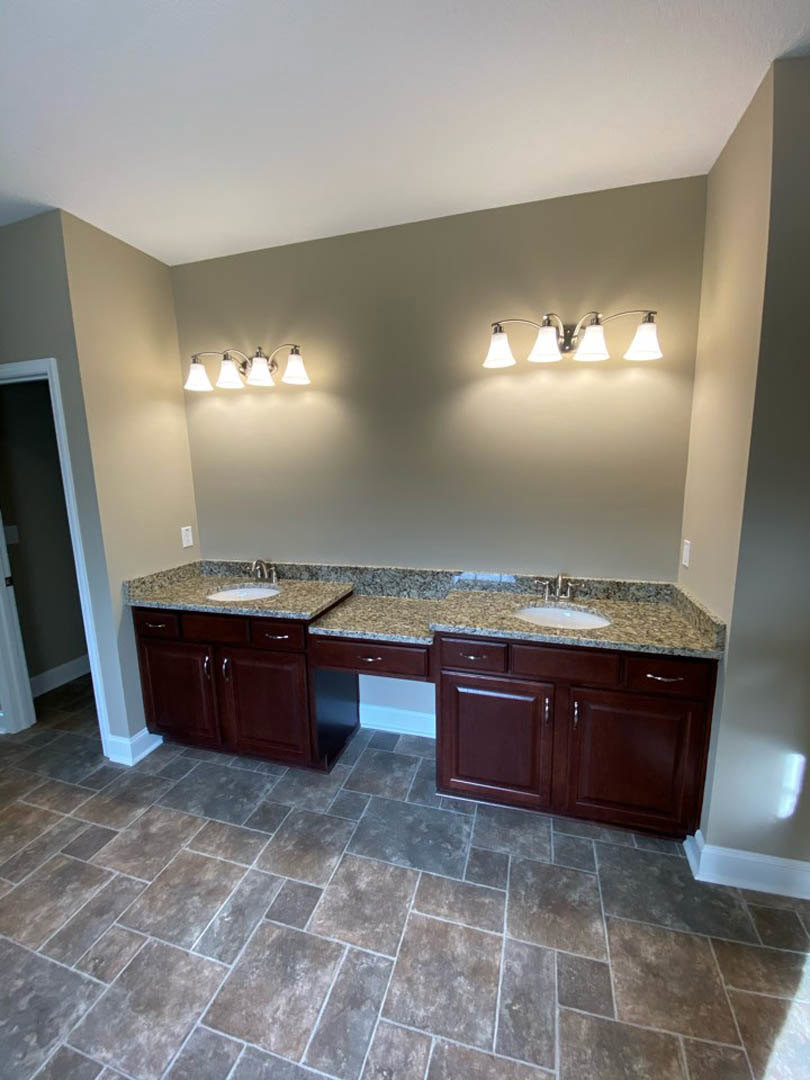 Bathroom with double sinks, polished marble countertops, white tile floor, dark wood cabinetry, and chrome faucets beneath a three-light fixture.