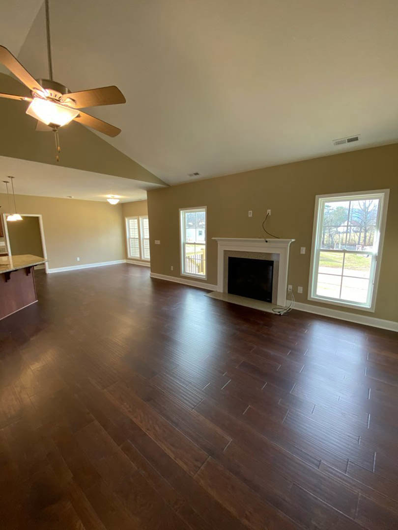 Living room with hardwood flooring, central fireplace with white mantel, ceiling fan, large windows with white frames overlooking yard, flat screen TV mounted on wall