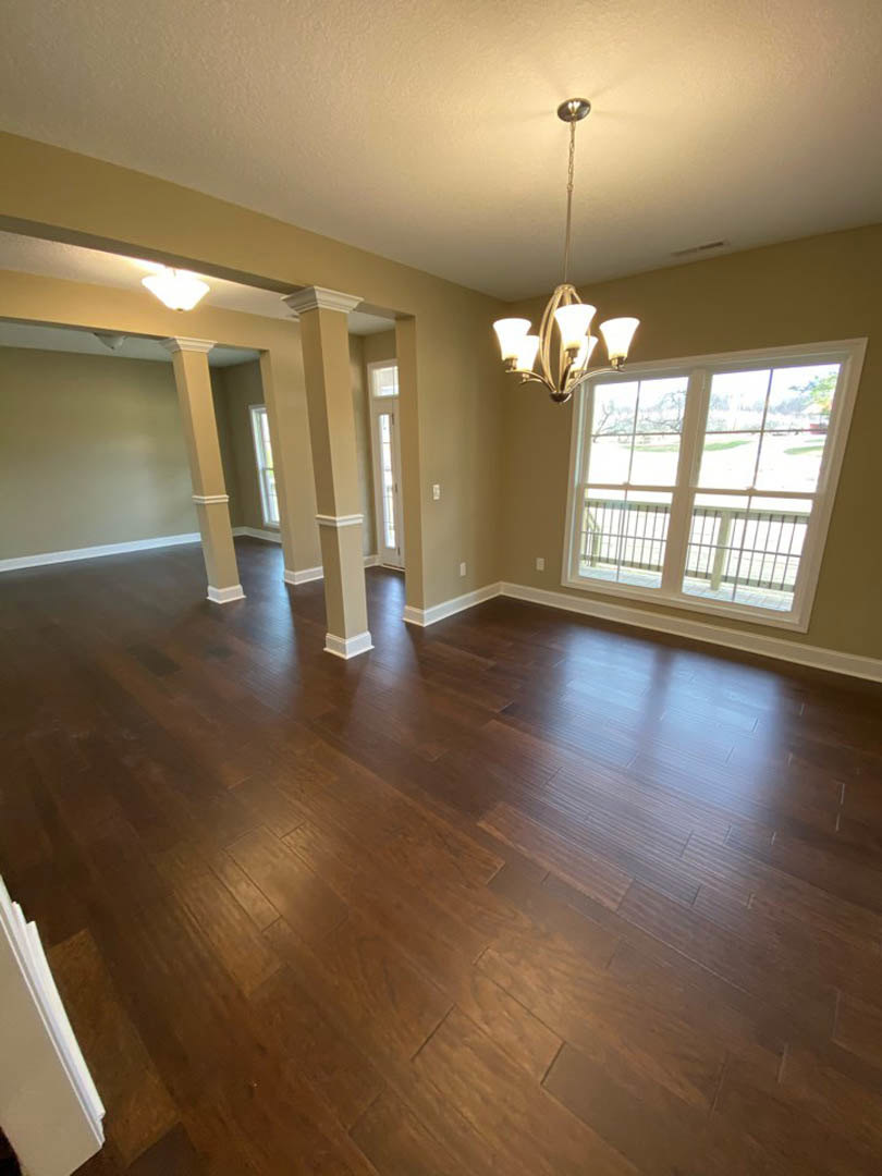 Open living room with dark hardwood floors, white pillars, large window with decorative railing, and ornate chandelier hanging from a white ceiling.
