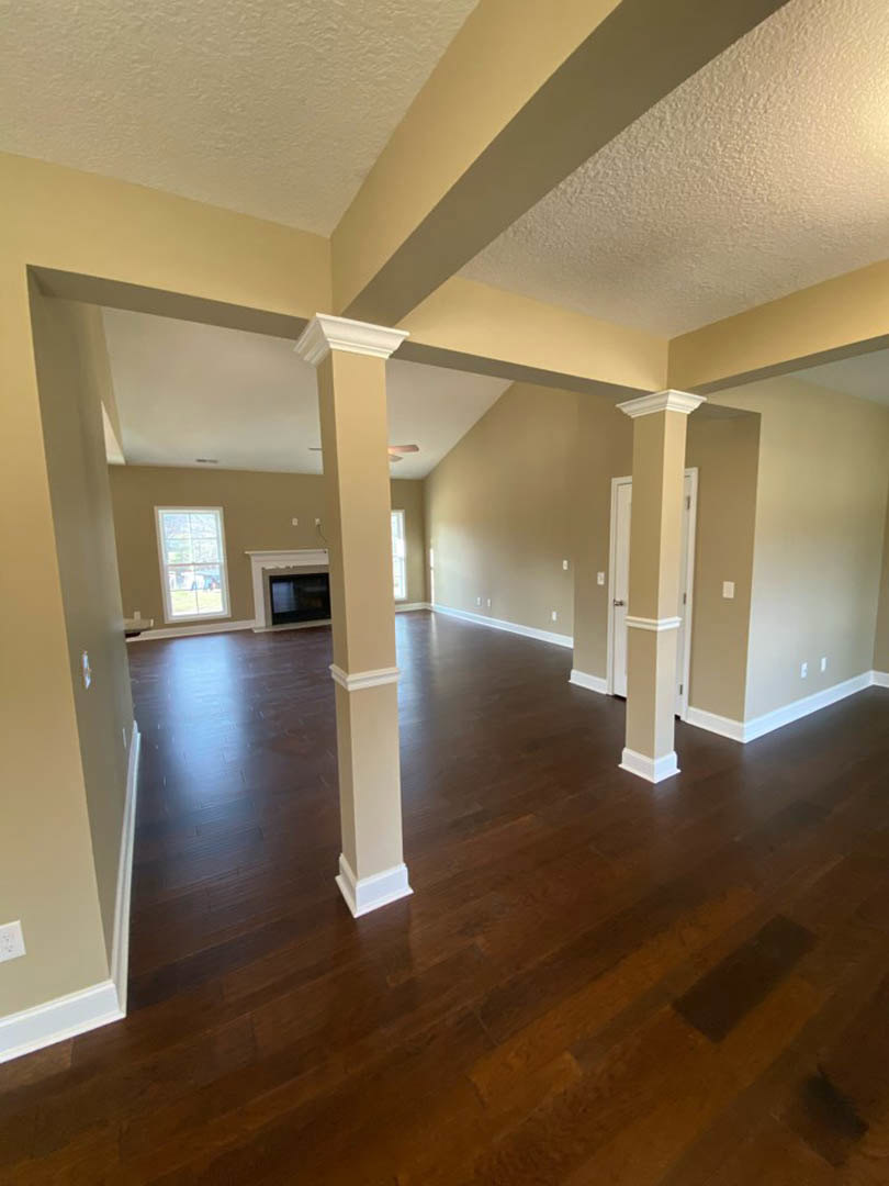 Open living room with white columns, hardwood flooring, white-framed window, and fireplace against a neutral wall.
