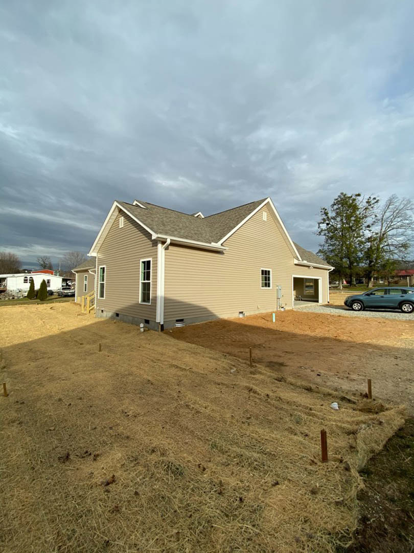 Two-story custom home with white siding, large windows, and a gray shingle roof; parked sedan in driveway, mature tree and patchy lawn in front, overcast sky above