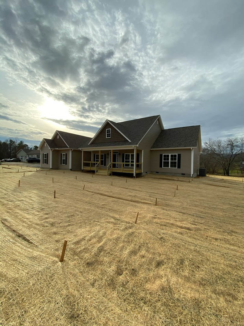 Two-story house with multi-pane windows, covered front porch, fenced grassy yard, surrounded by mature trees under a cloudy sky