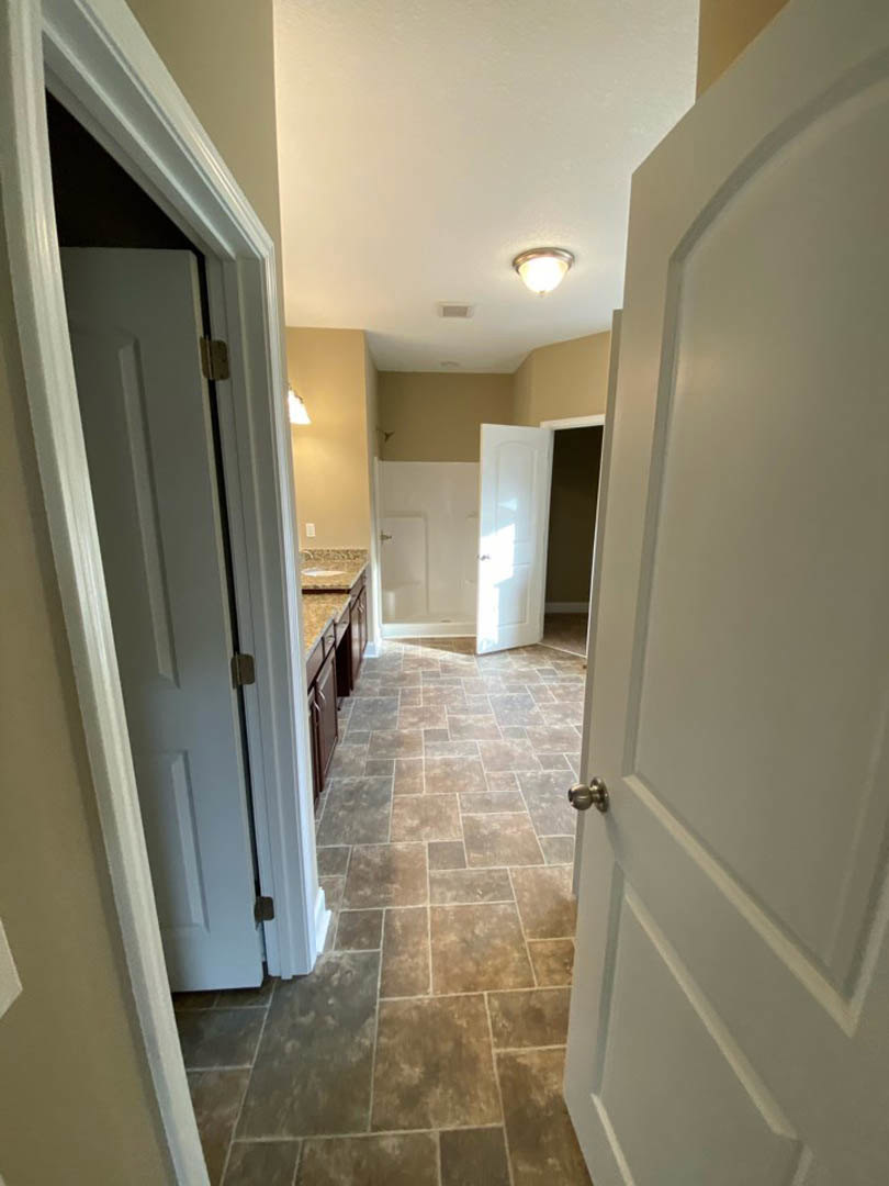 Hallway with white paneled doors, brown ceramic tile flooring, white plaster walls, ceiling light fixture, and open door letting in natural light