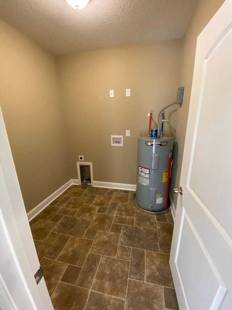 Large grey water heater against a white wall, brown tile flooring, white door with black handle, indoor utility room