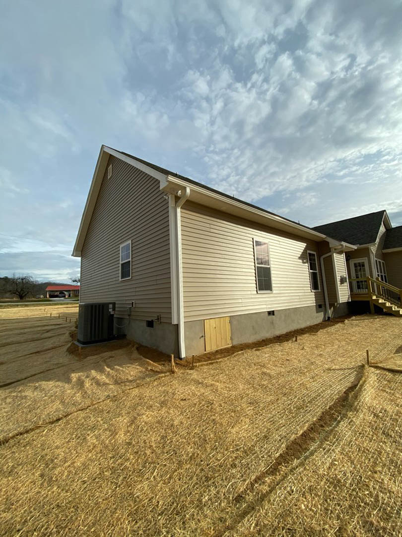 Framed house under construction with exposed wood panels, surrounded by a field of hay under a partly cloudy sky