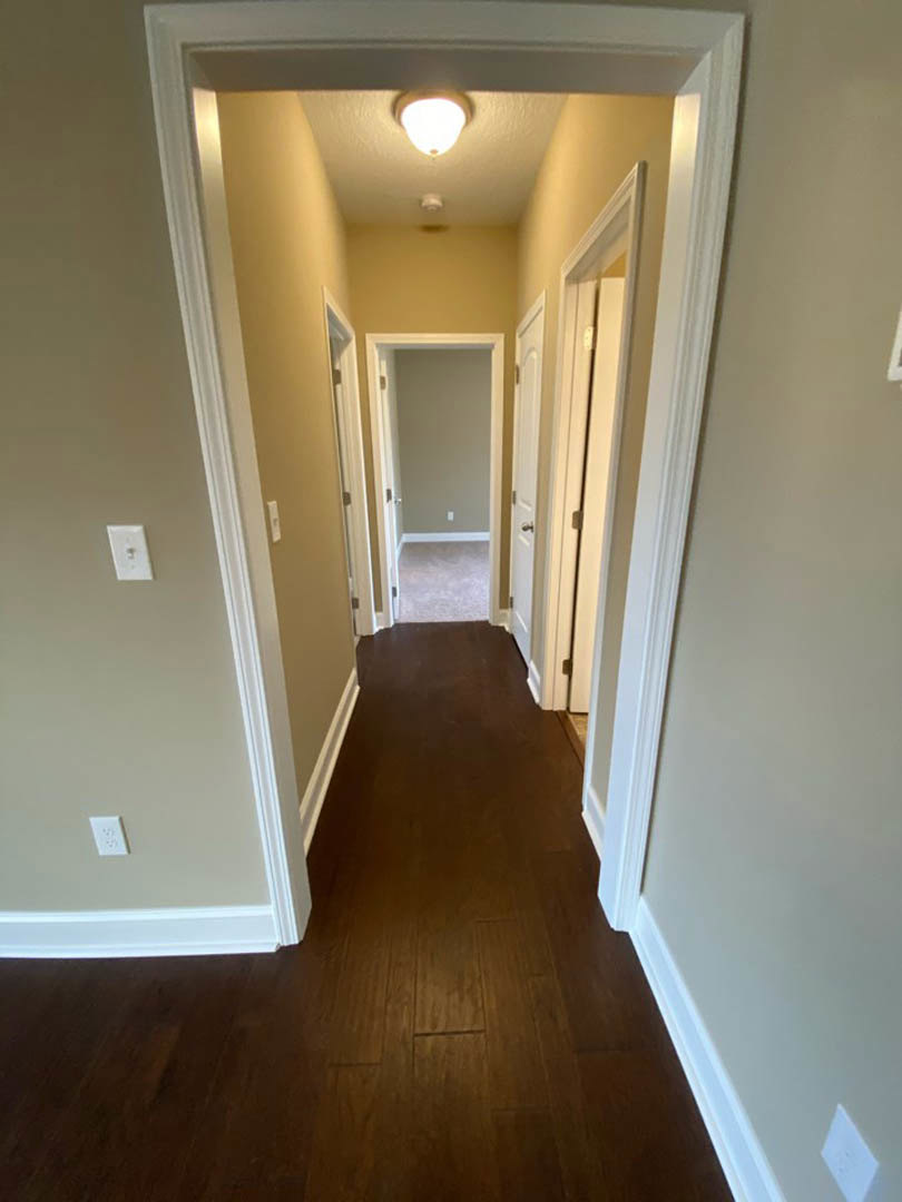 Hallway with dark wood laminate floors, white walls, crown molding, illuminated ceiling light fixture, white door, and electrical outlet