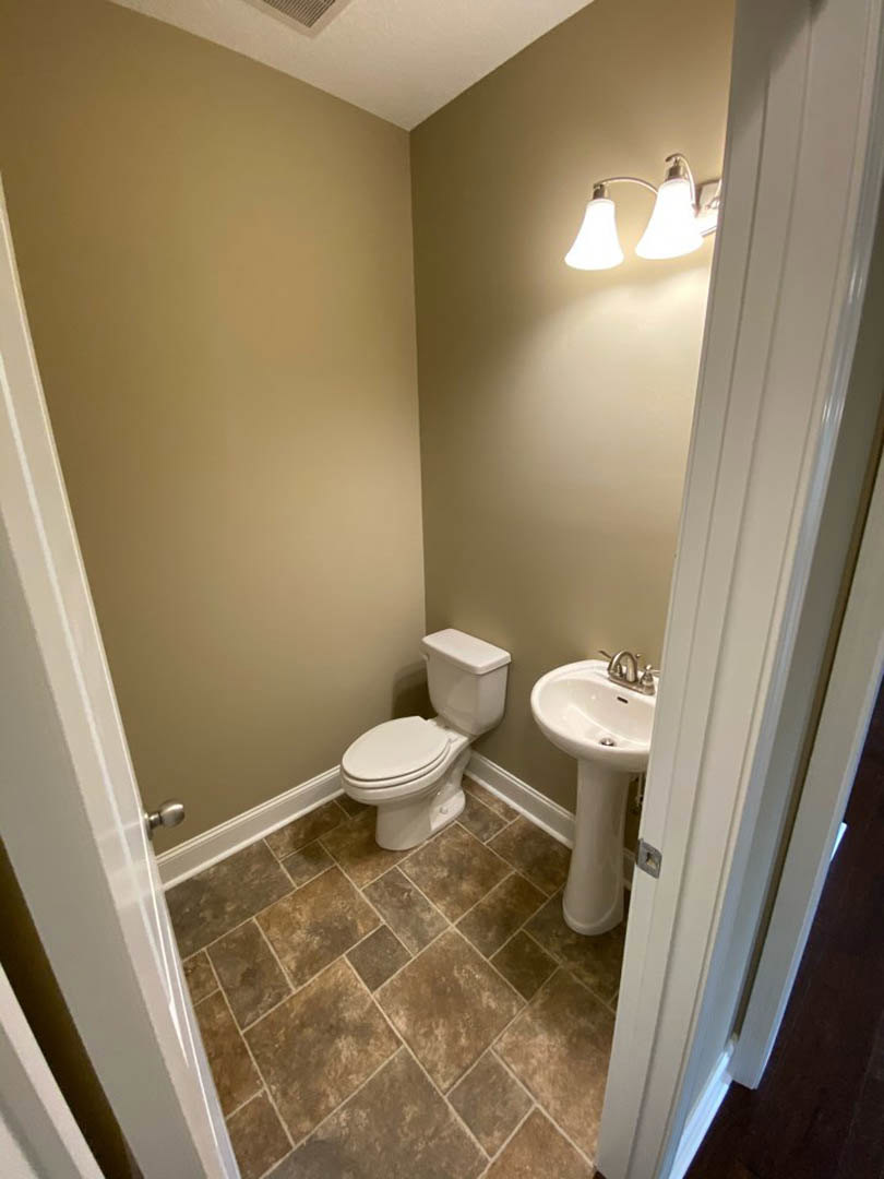 White pedestal sink and toilet in a bathroom with brown tile floor, wall-mounted mirror, and two-light fixture above sink.
