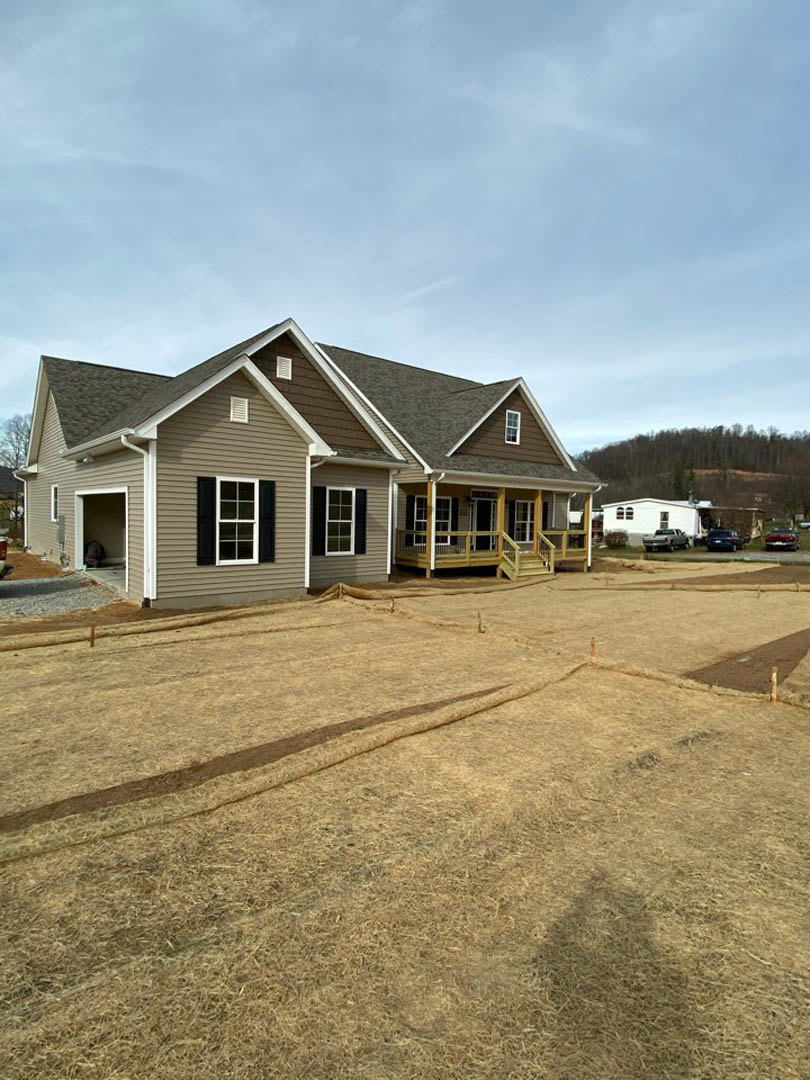 Two-story home with white siding, black shuttered windows, covered front porch, expansive grassy yard bordered by a wooden fence, mature trees, and a person standing in the doorway
