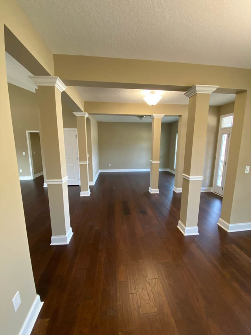 Hardwood floor with white pillars, tan walls, white paneled door, ceiling light fixture, and window letting in natural light