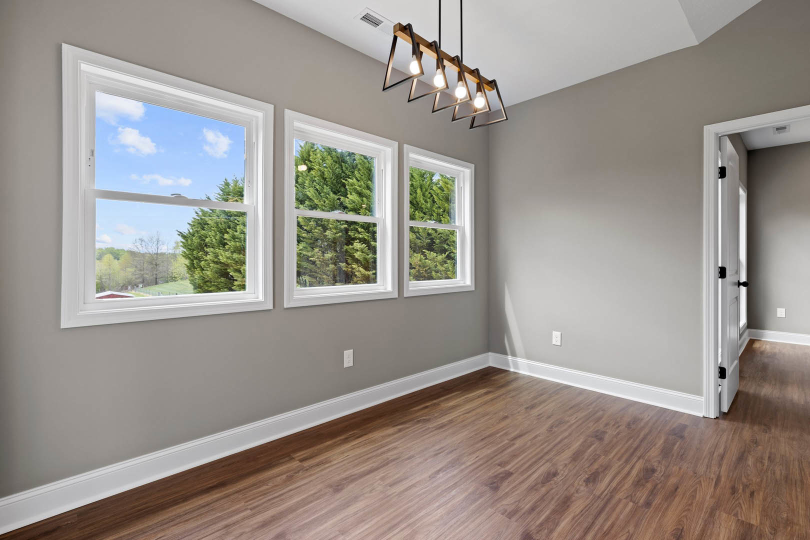 Spacious room with two large windows overlooking grassy field and trees, wood flooring with white baseboards, modern ceiling light fixture with exposed bulbs