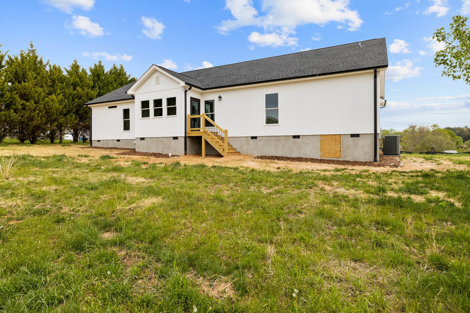Two-story house with white-framed windows, wooden staircase leading to entry, surrounded by green grass yard and mature trees under partly cloudy sky
