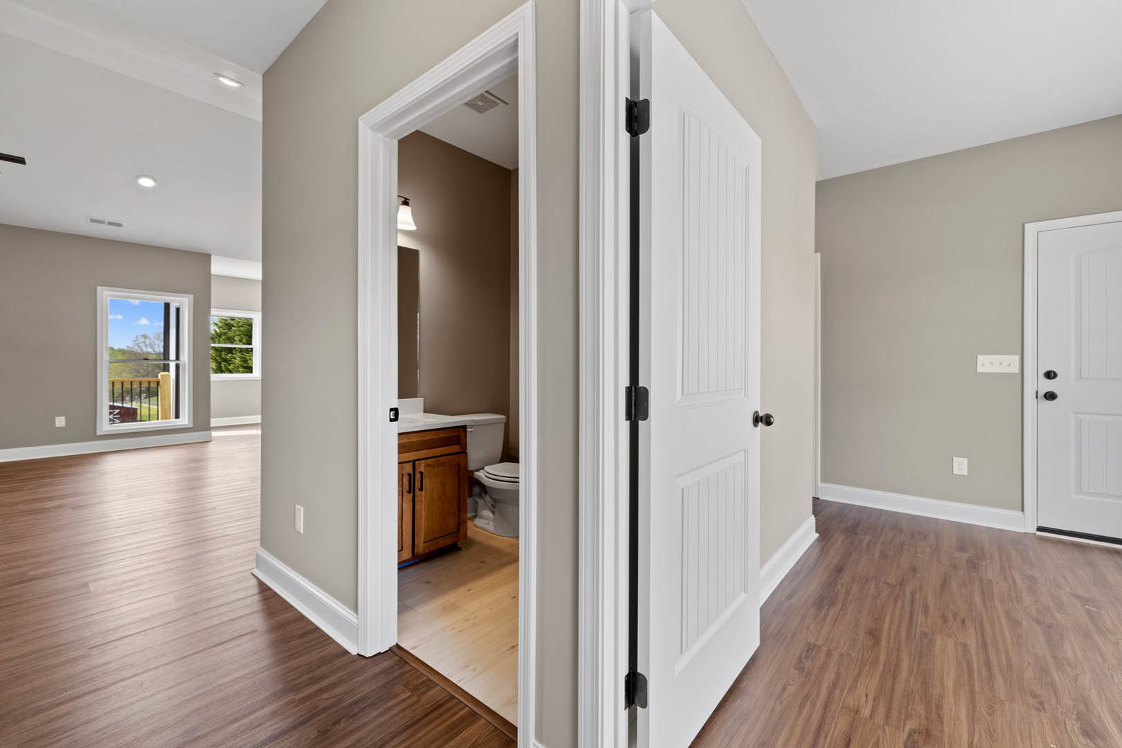 Hallway with open white door, hardwood floor, white walls, wooden cabinet, and window showing outdoor view