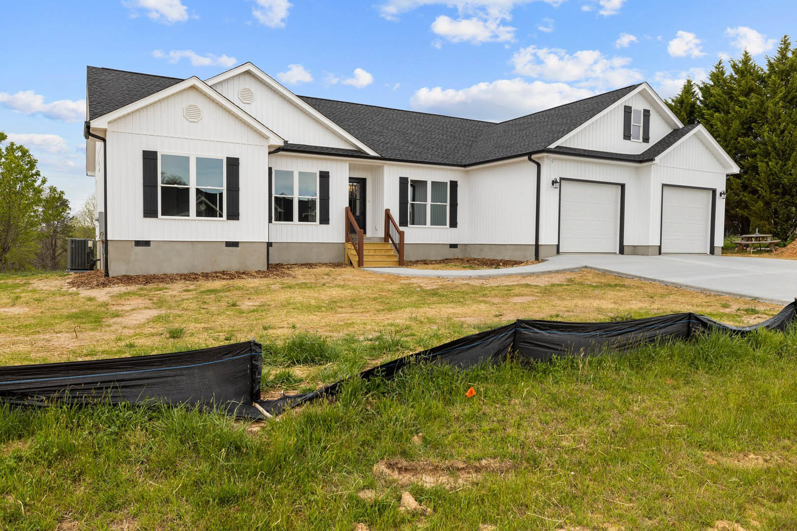 White house with black metal fence, wooden staircase leading to entry, black tarp on lawn, large window overlooking tree, white garage door framed in black.